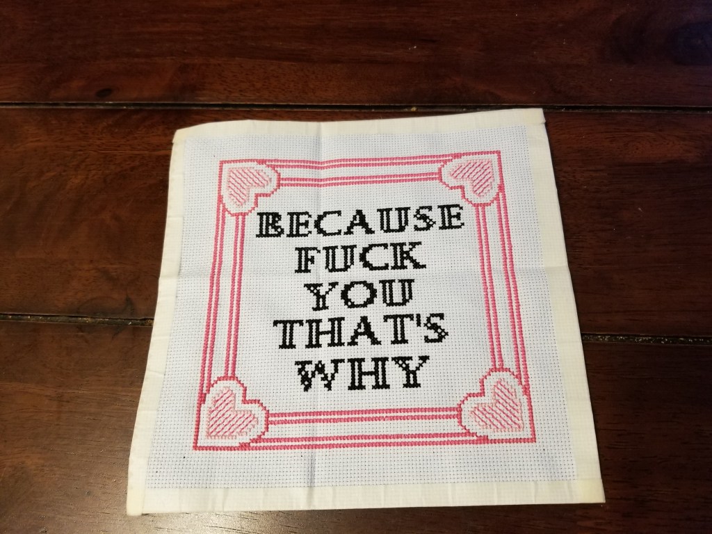Because Cross Stitch. A homey looking cross stitch with a pretty pink border (three different pinks!) and embedded heart shapes that surrounds the message “Because fuck you that’s why” in traditional cross stitch sampler font. The cloth itself is approximately 7.5” by 7.5”, square, and the design is approximately 5.5” x 5.5”.