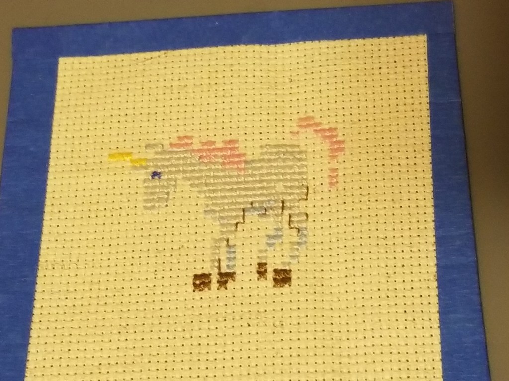 Item: Unicorn Cross Stitch. A silvery white unicorn with a yellow horn, a pink mane and a pink tail. The cloth itself is approximately 4.25” by 4.25”, square, and the design is approximately 2” x 2.5”.