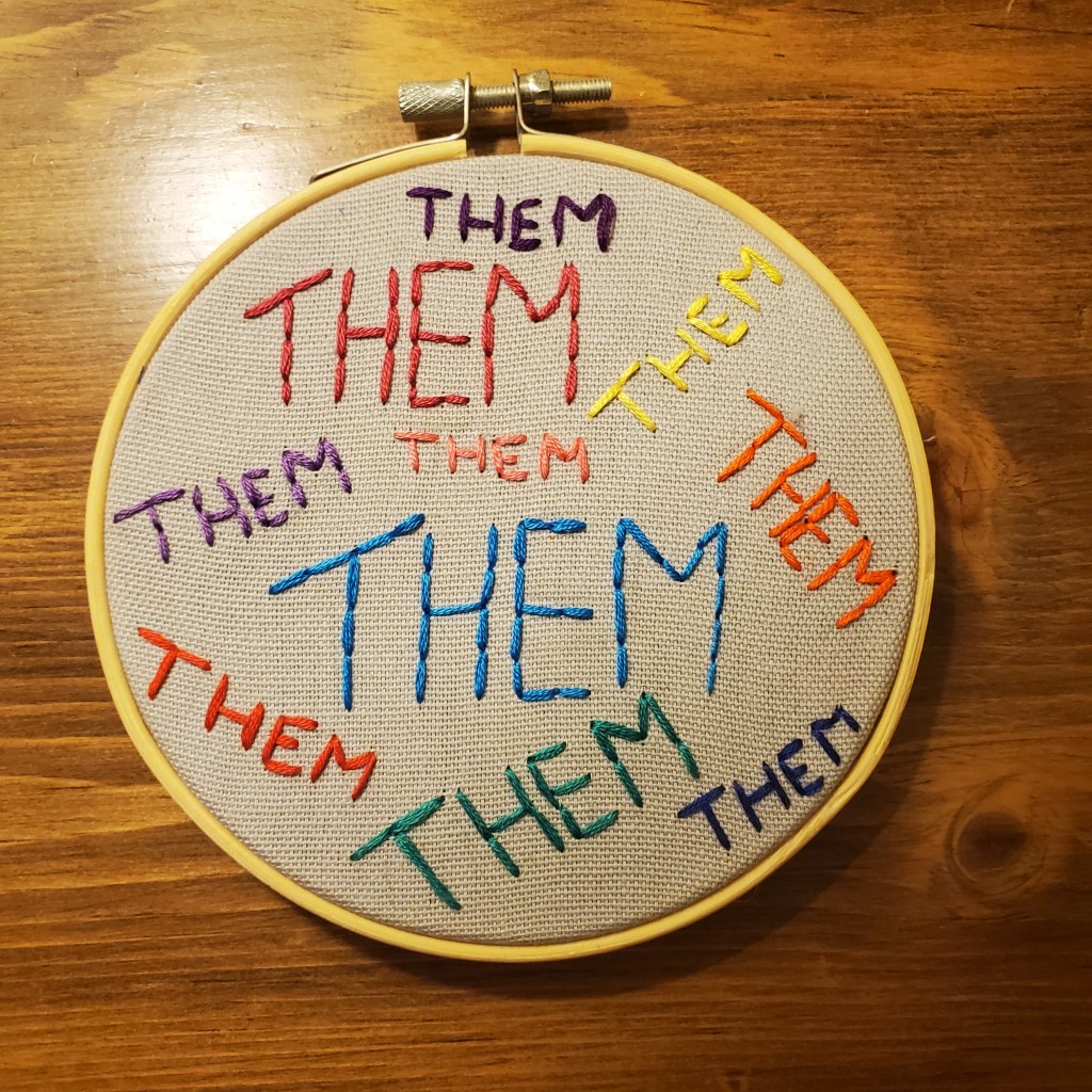 Item: Them Pronoun Embroidery. A five-inch circular embroidery on grey fabric with “Them” written in neon colours.
